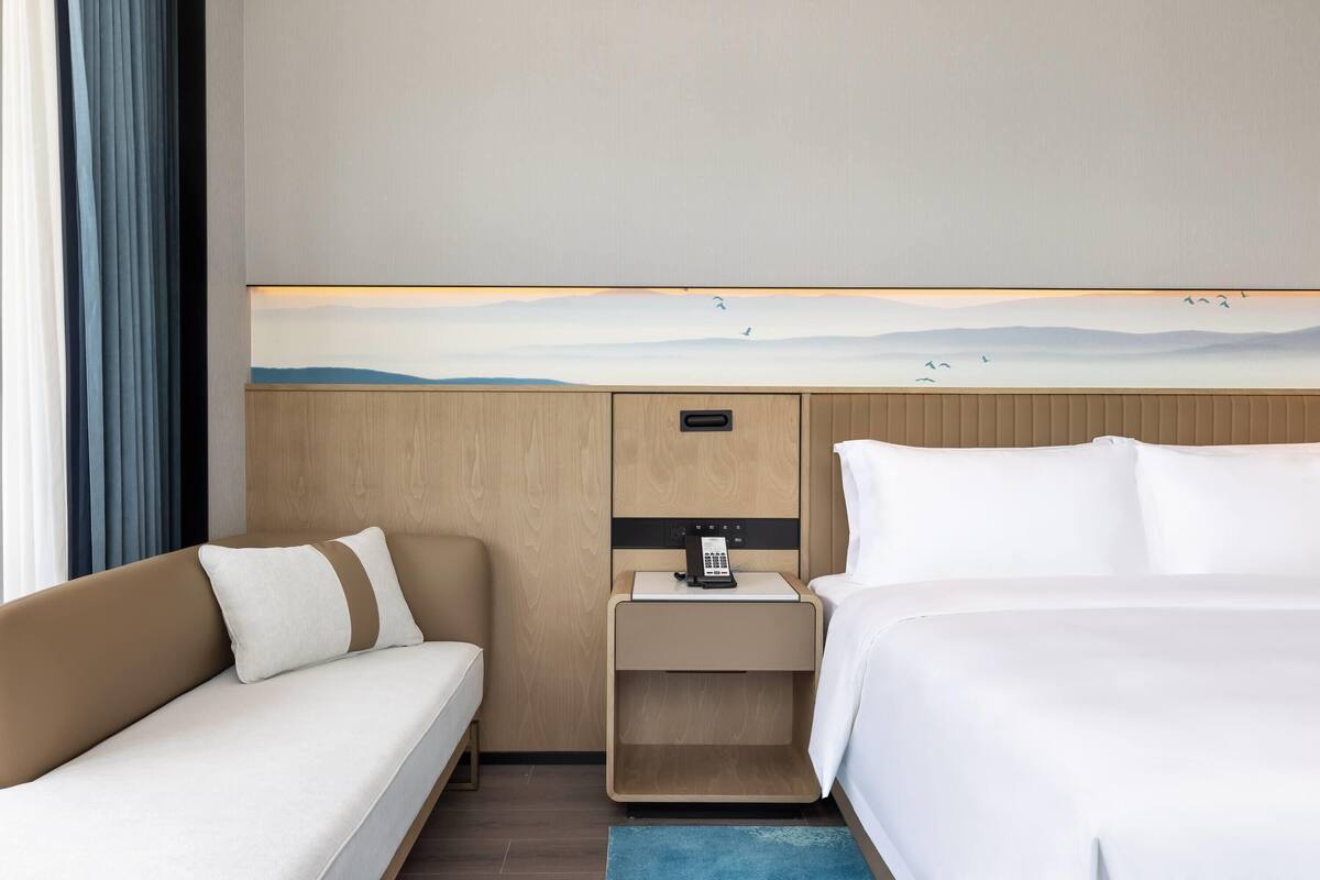 A hotel room with a bed, a sofa, a nightstand with a phone, and a wall-mounted artwork.