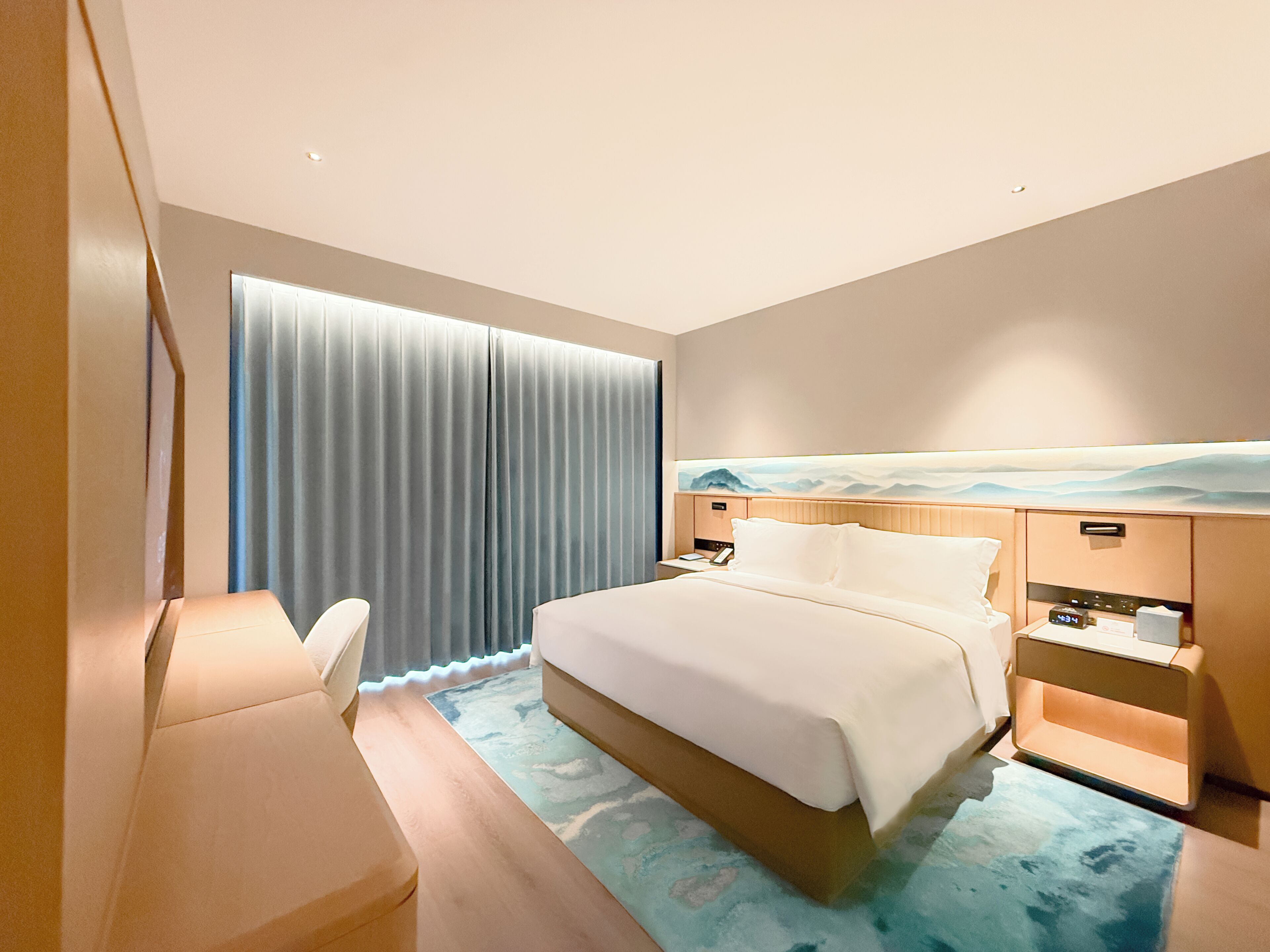 A modern hotel room with a large bed, bedside tables, a desk, and a window with curtains.