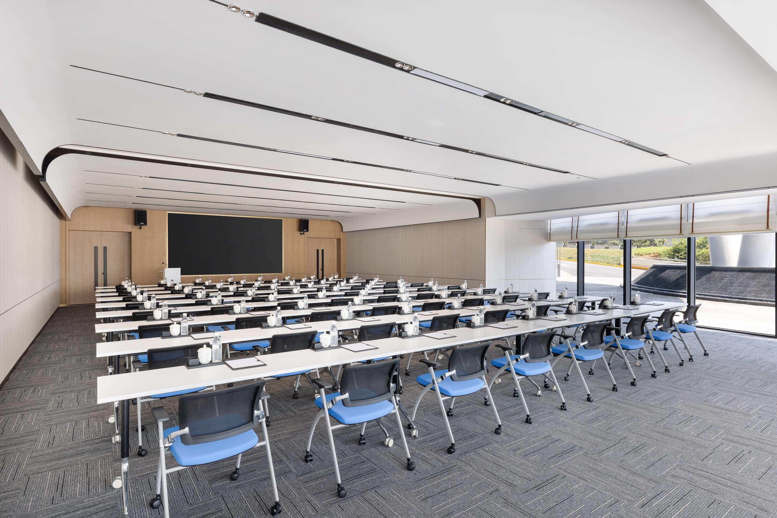 A conference room with rows of tables and chairs, a large screen, and a view of the outdoors.