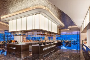 A modern hotel lobby with a central bar area, seating arrangements, and a view of the cityscape through large windows.