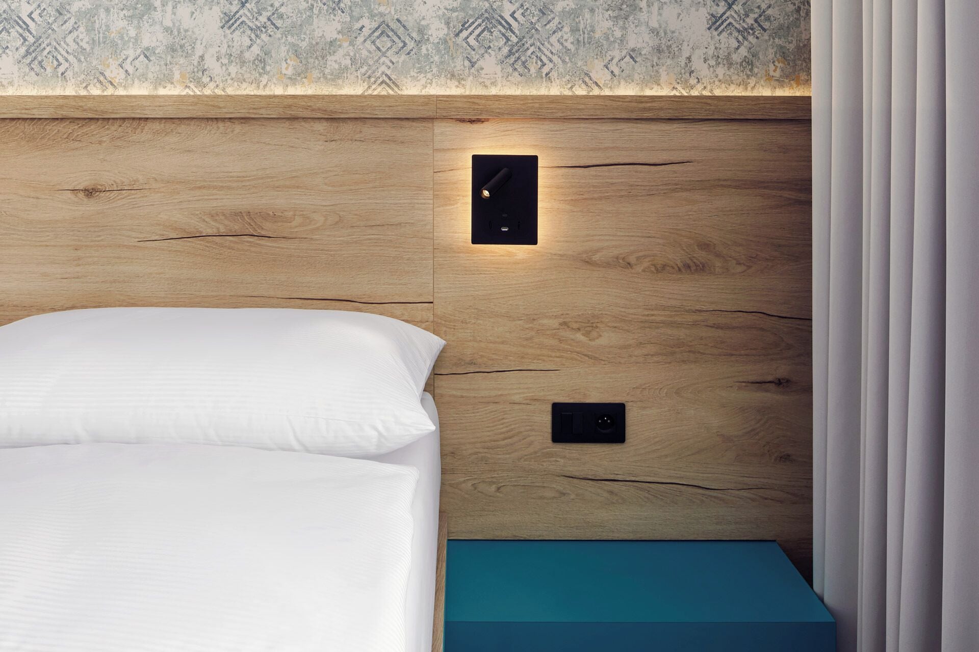 A neatly made bed with white linens, a wooden headboard with built-in lighting, and a teal bedside table.
