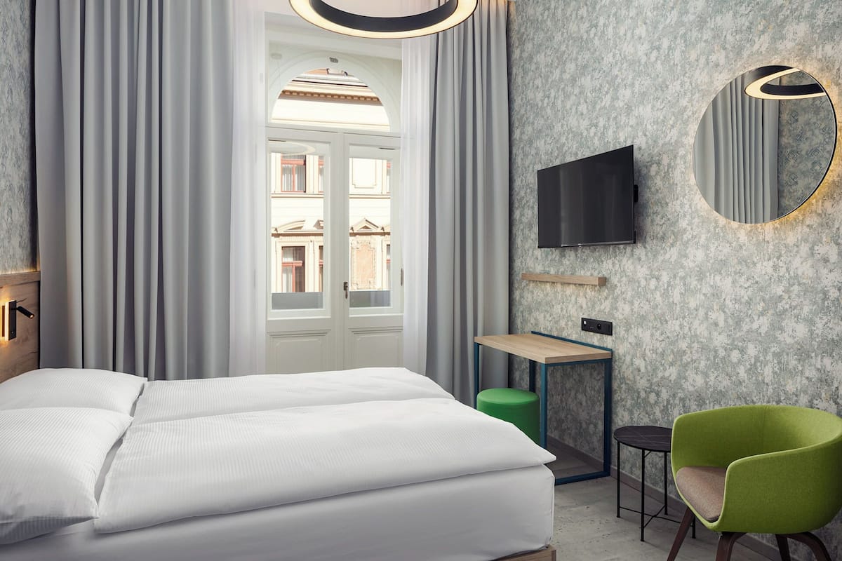 Une chambre d’hôtel avec un lit, un téléviseur fixé au mur, une petite table, une chaise verte et une vue sur les bâtiments par la fenêtre.