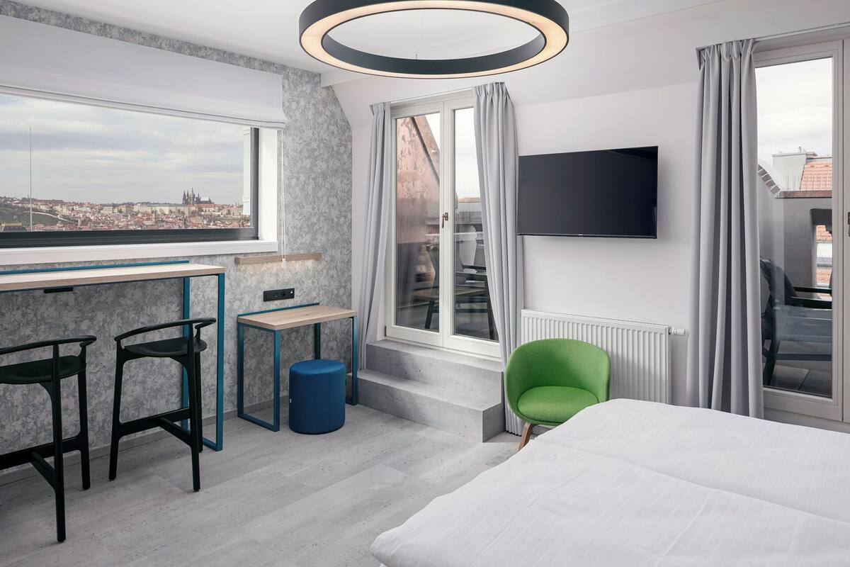 Une chambre d’hôtel moderne avec vue sur la ville, un bureau, deux chaises, un fauteuil vert et un lit.