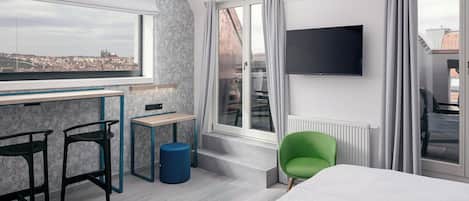 A modern hotel room with a city view, a desk, two chairs, a green armchair, and a bed.