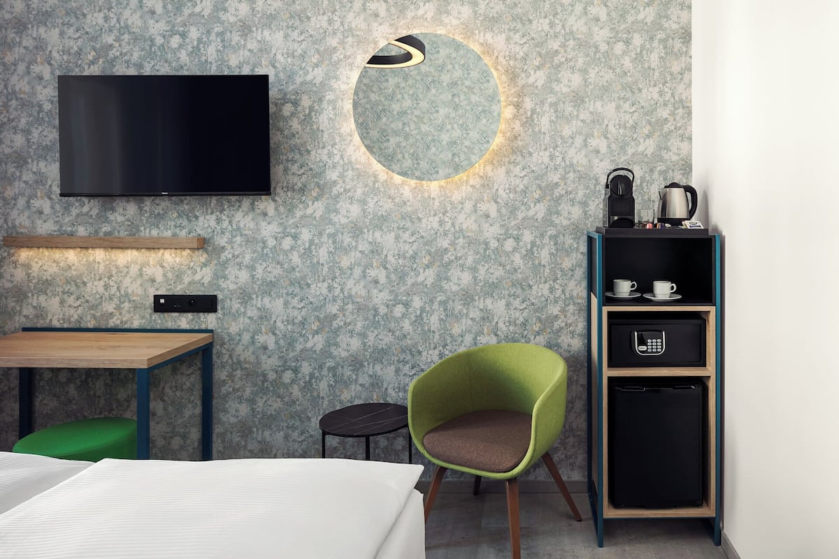 Une chambre d’hôtel moderne équipée d’un téléviseur à écran plat, d’un luminaire rond, d’une petite table ronde, d’un fauteuil vert, d’un minibar et d’un lit.