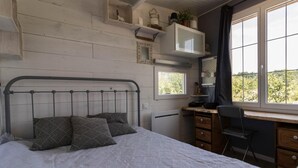 Un dormitorio con cama, escritorio y silla. Se ve vegetación por la ventana.