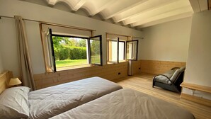 A bedroom with two beds, large windows, a wooden floor, and a view of greenery outside.