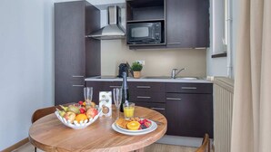 Eine kleine Küche mit dunklen Schränken, einer Mikrowelle und einem Spülbecken. Im Vordergrund steht ein Tisch mit Obst und Saft.