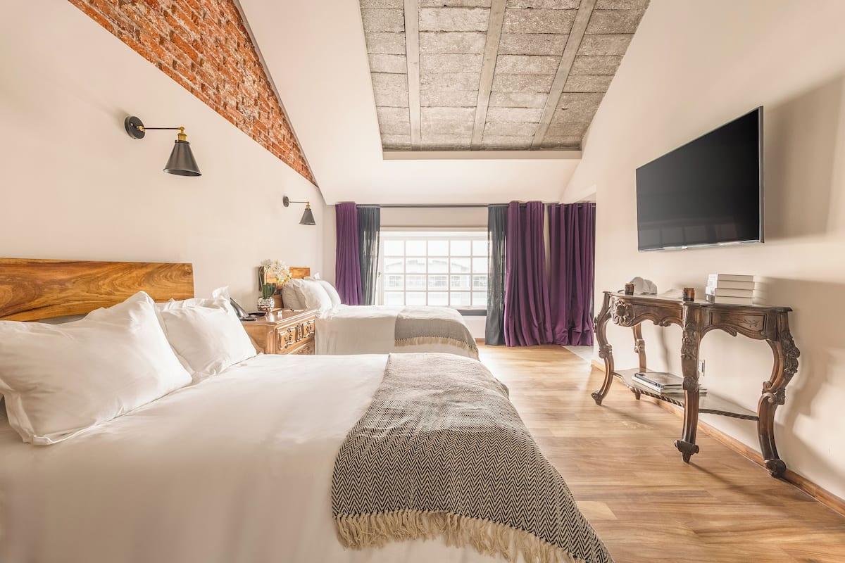 Une chambre d’hôtel avec un grand lit, une tête de lit en bois, un téléviseur à écran plat fixé au mur et une fenêtre ornée de rideaux violets.