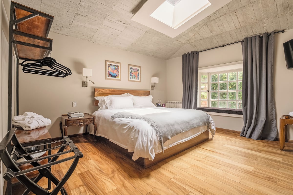 Une chambre avec un plancher en bois, un lit avec une literie blanche, une tête de lit en bois, une table de chevet avec une lampe, un porte-manteau mural et une fenêtre avec des rideaux.