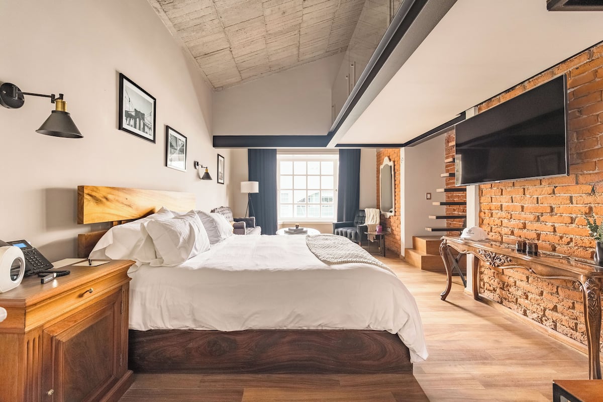 Une chambre moderne avec un grand lit, une tête de lit en bois, un téléviseur à écran plat fixé à un mur en briques et un bureau avec une lampe.