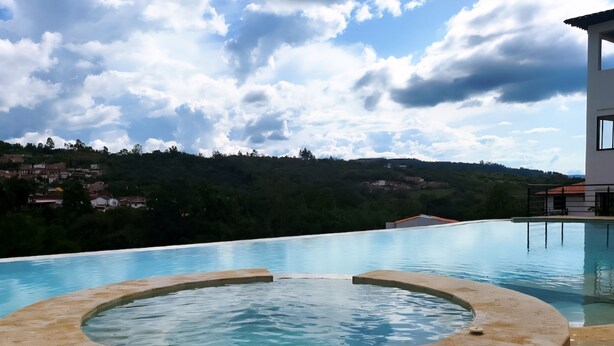 Une piscine avec un jacuzzi au centre, entourée d’une terrasse en pierre, et offrant une vue sur une colline parsemée de maisons.