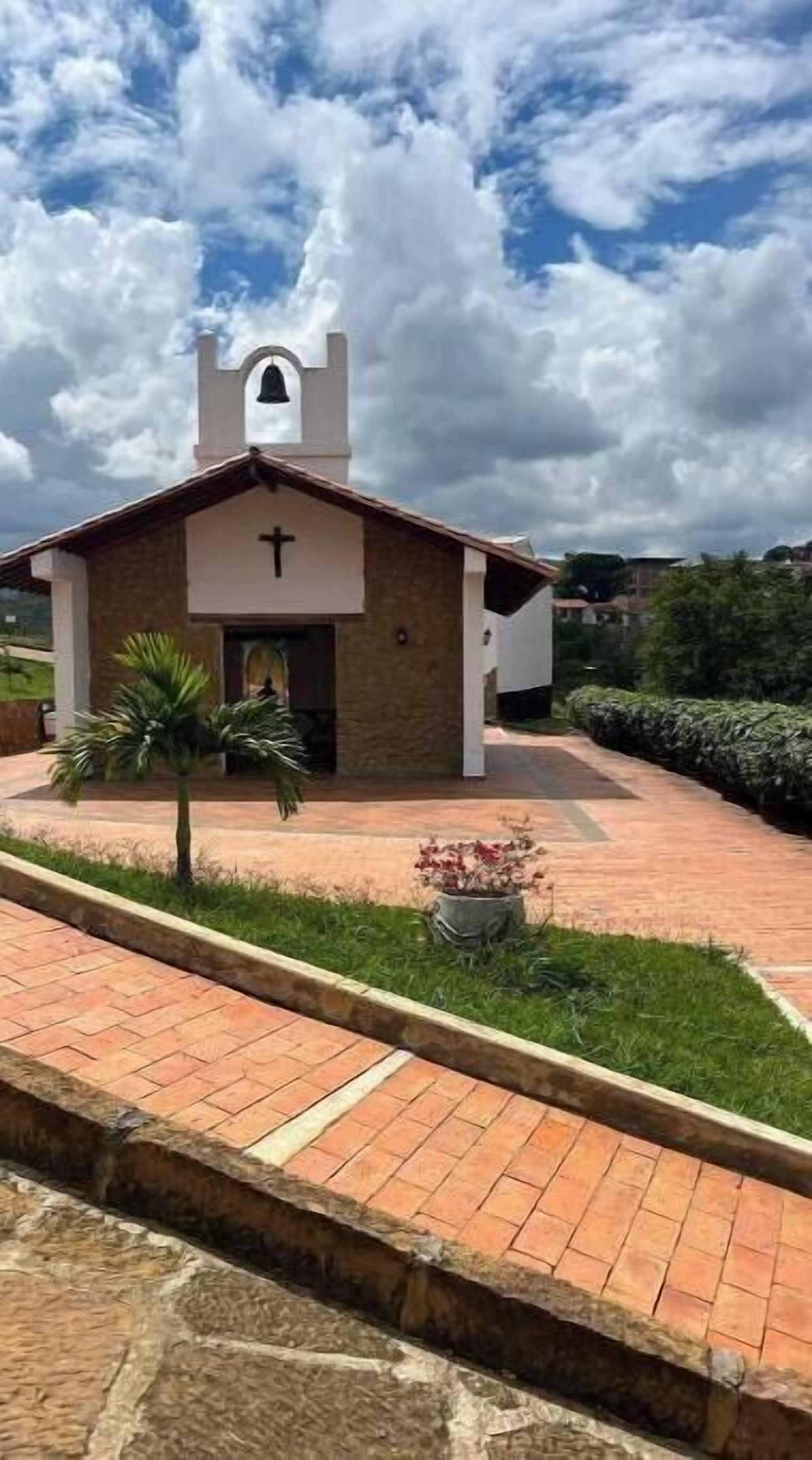 Uma pequena igreja com um campanário, uma cruz na fachada e um caminho de pedras que a leva até ela.