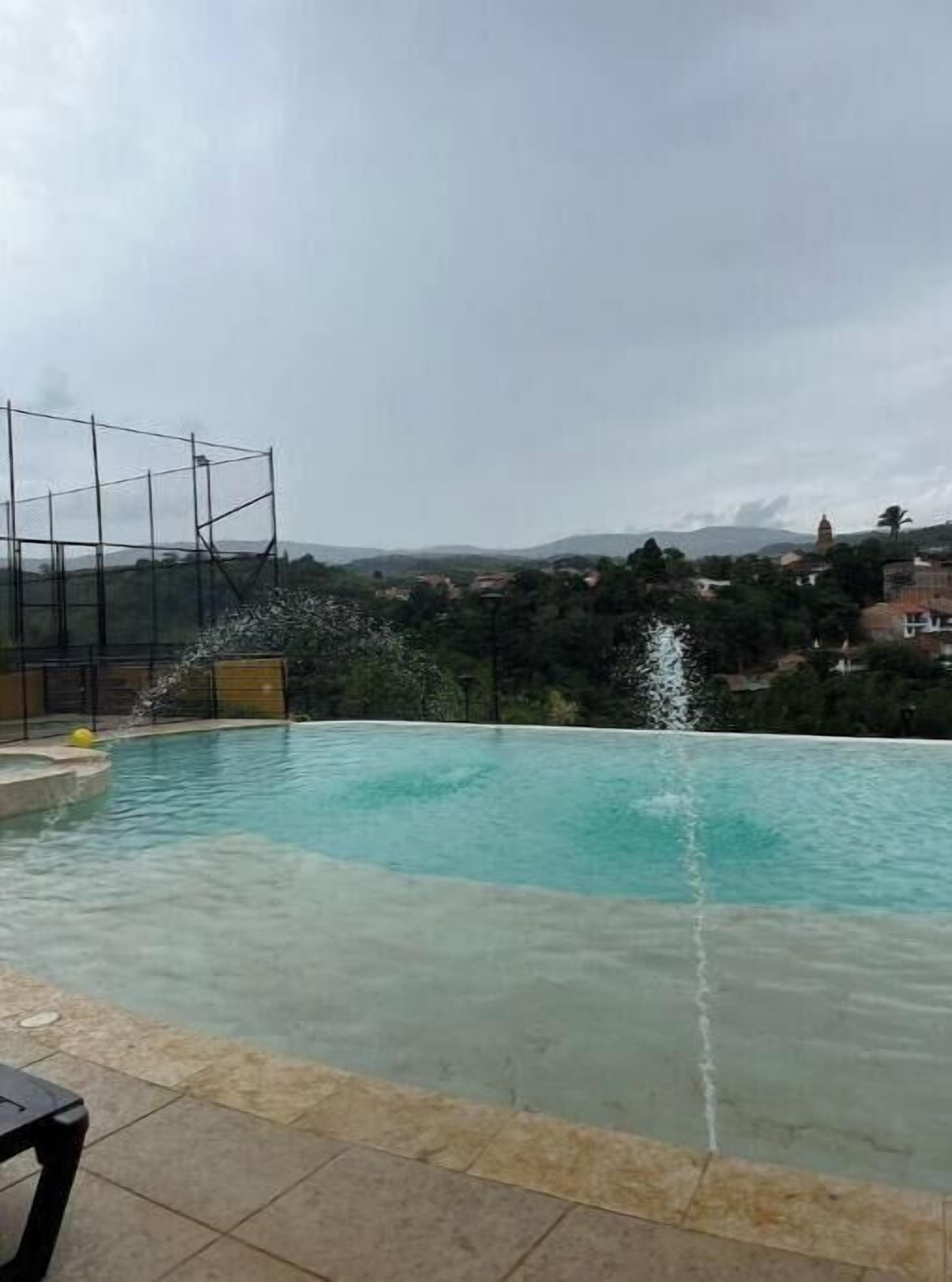 A swimming pool with a water feature, surrounded by a fence and overlooking a hilly landscape.