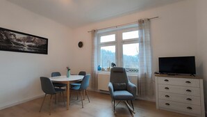 A room with a round dining table, four chairs, a television on a wooden cabinet, a window with curtains, and a painting on the wall.