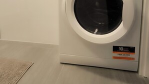 A front-loading washing machine in a room with a light-colored floor and a small shelf above it.