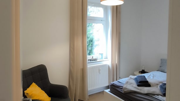 A bedroom with a bed, a chair with a pillow, a hanging light fixture, and a window with curtains.