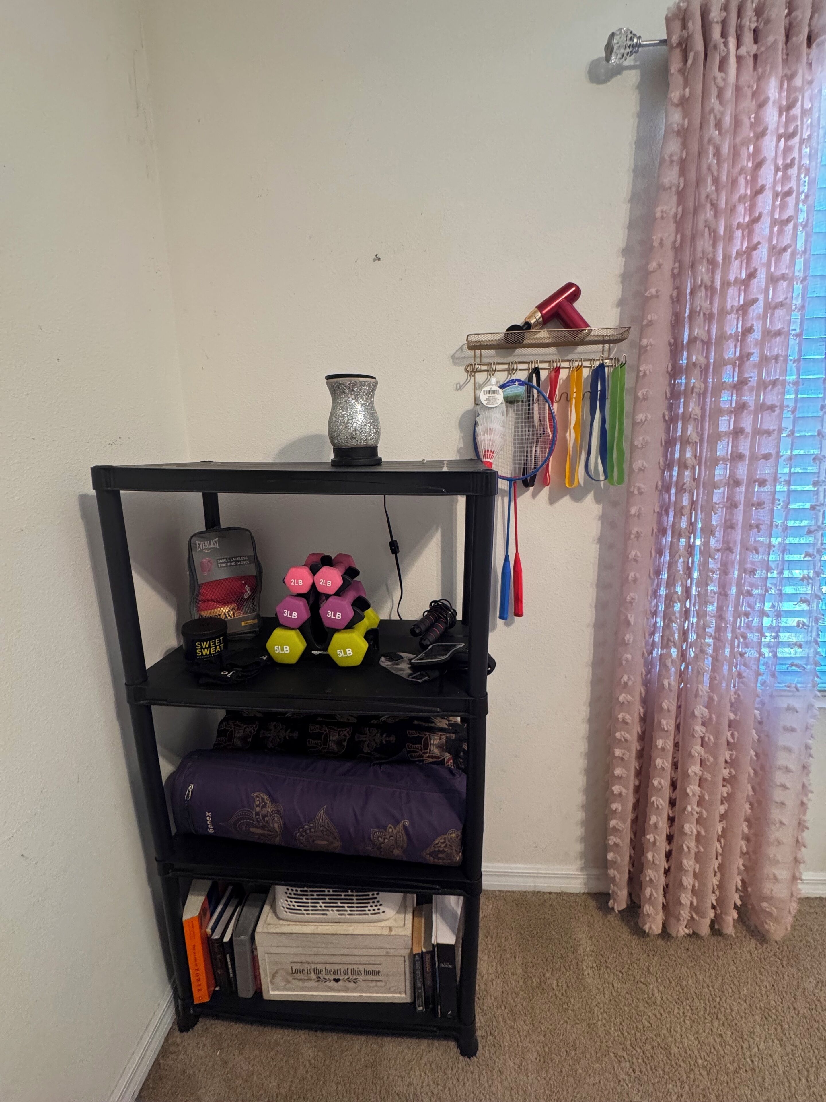 A room with a black shelf containing dumbbells, a sweet sweat candle, and a decorative item. There is a window with pink curtains and a shelf with badminton rackets.