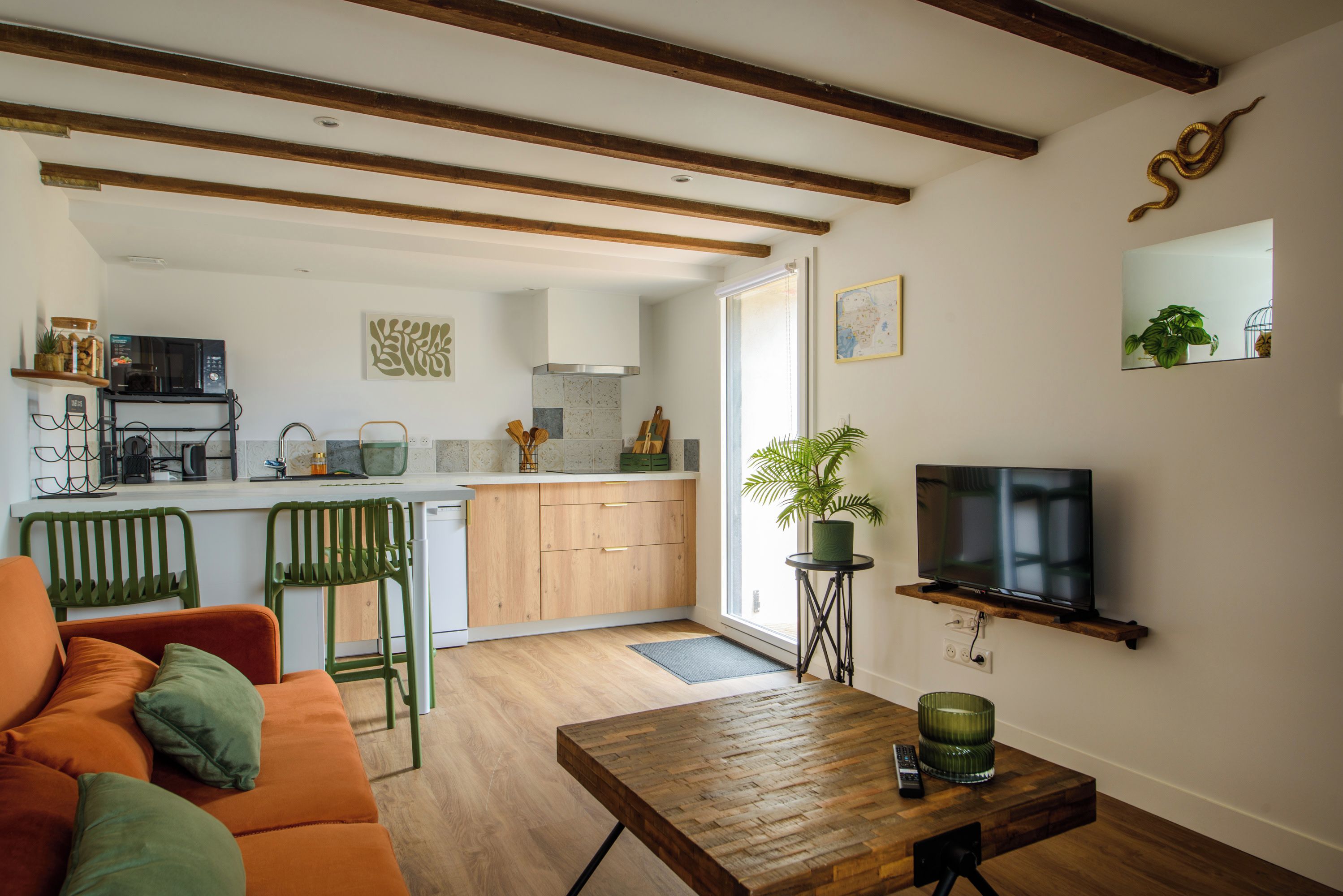 A modern living room with a wooden coffee table, a sofa, and a television. There is a kitchen in the background with a microwave and a sink.