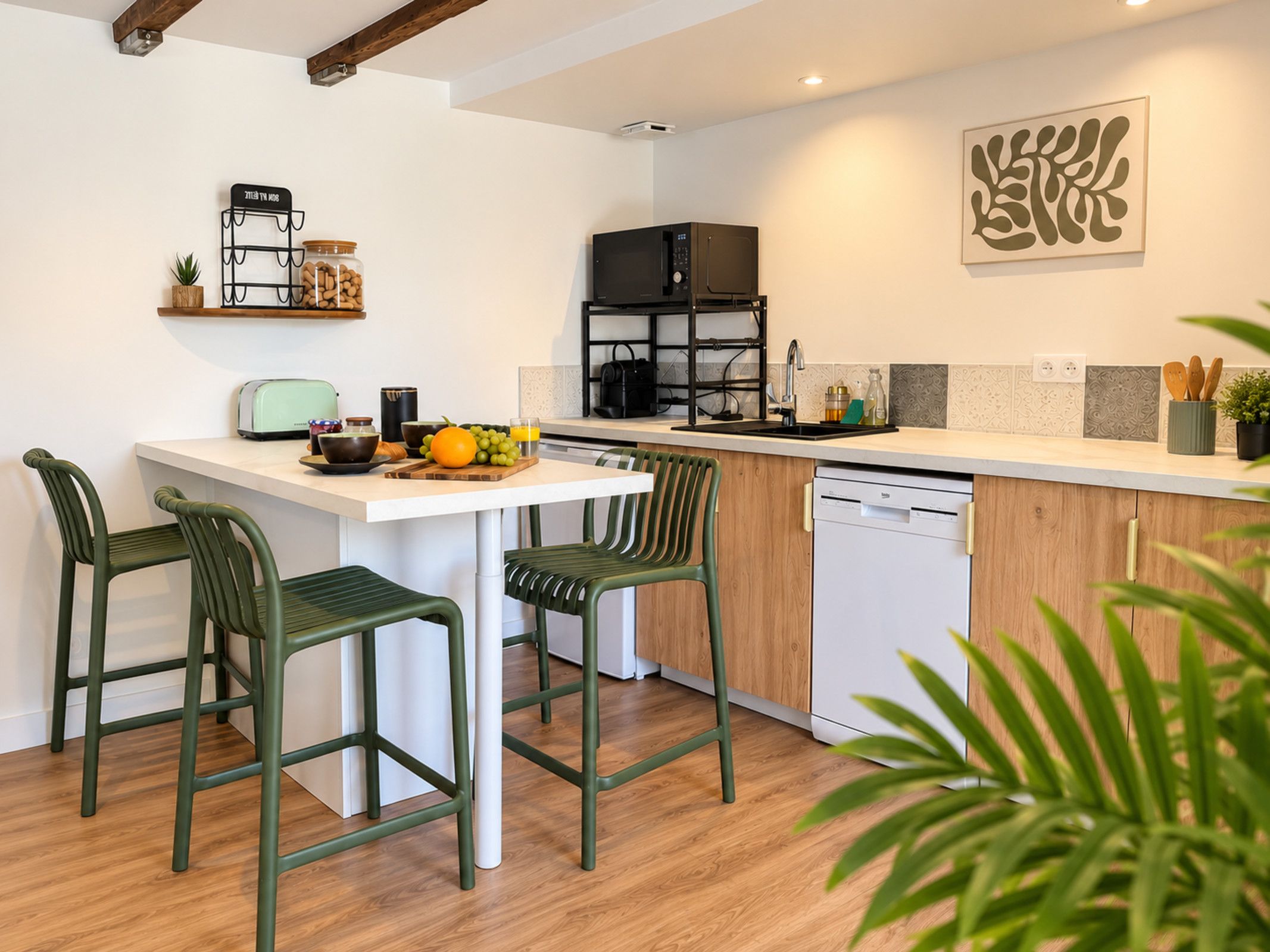 Une cuisine moderne avec un plan de travail blanc, deux chaises vertes, un micro-ondes noir et un lave-vaisselle.