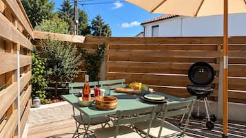 Um pátio externo com mesa de madeira, cadeiras, churrasqueira e um grande guarda-sol.