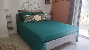 A bed with teal bedding and patterned pillows, a wooden headboard, a small bedside table with a lamp, and a view of the outdoors through a window. - Room N°2 Privée & SDB｜Villa au Calme & Parking - Chez Benny (Nice)