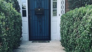 Une porte d’entrée noire avec une boîte aux lettres, bordée de haies taillées, et un bâtiment en briques avec des ornements blancs et une plaque portant le numéro « 1000 ».