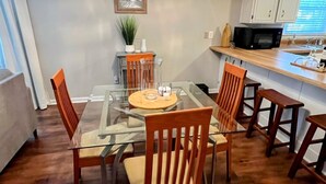 Un coin repas avec une table en verre et des chaises en bois, un luminaire suspendu, et une cuisine dotée de meubles blancs et d’un four à micro-ondes.