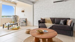 A modern living room with a dark sofa, a round wooden coffee table, and a dining table with chairs. There is a large window providing a view of the outdoors. - Lagoon view & Sunset ! Full unit 2BDR 2,5BTH (SAINT MARTIN)
