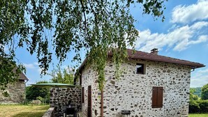 A rustic stone house with a tiled roof, a small window, and a door. There is a covered outdoor seating area with a table and chairs.