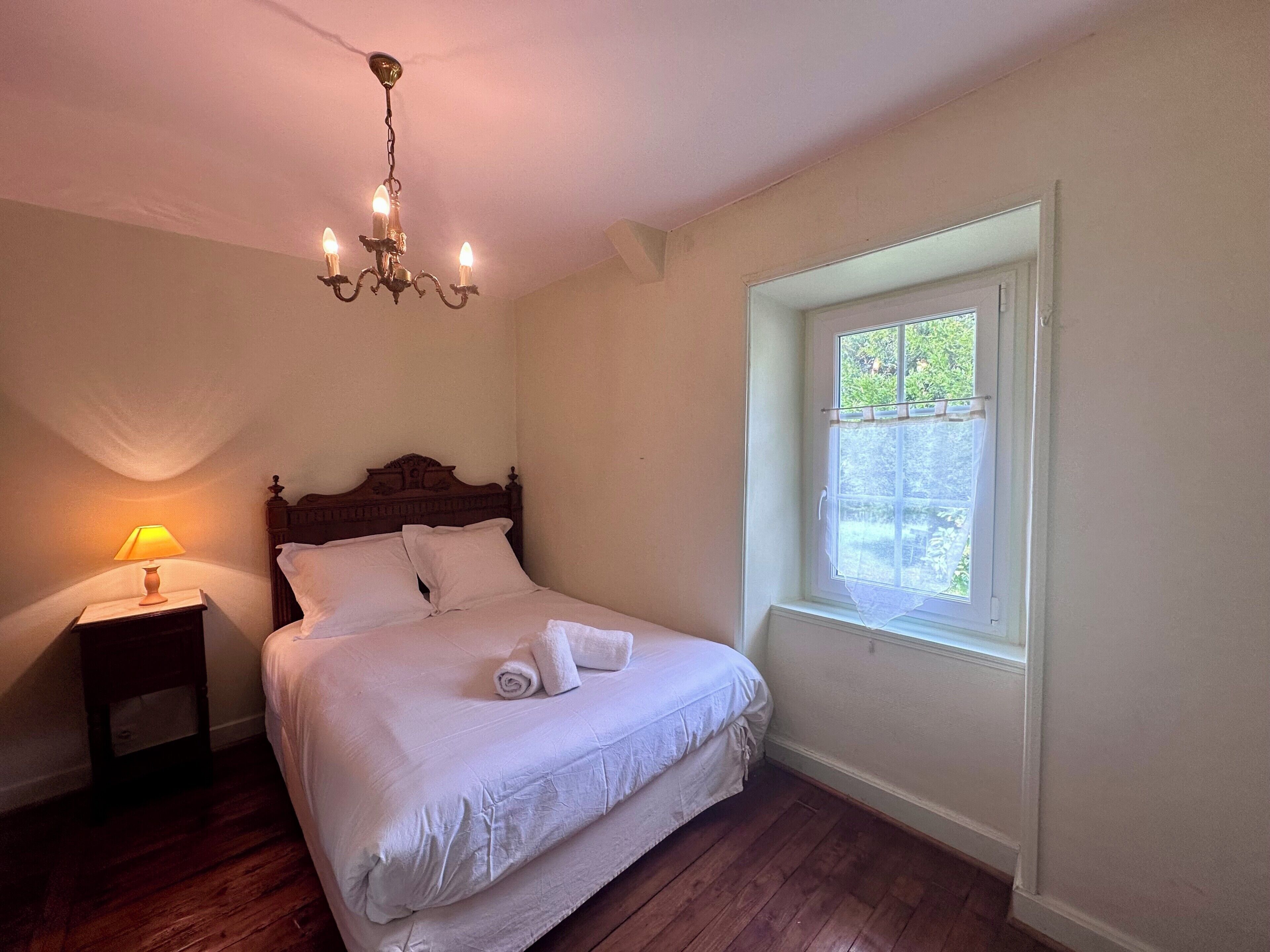 Un dormitorio con cama de madera, una mesita de noche con lámpara, una ventana con vistas a la vegetación y una araña.