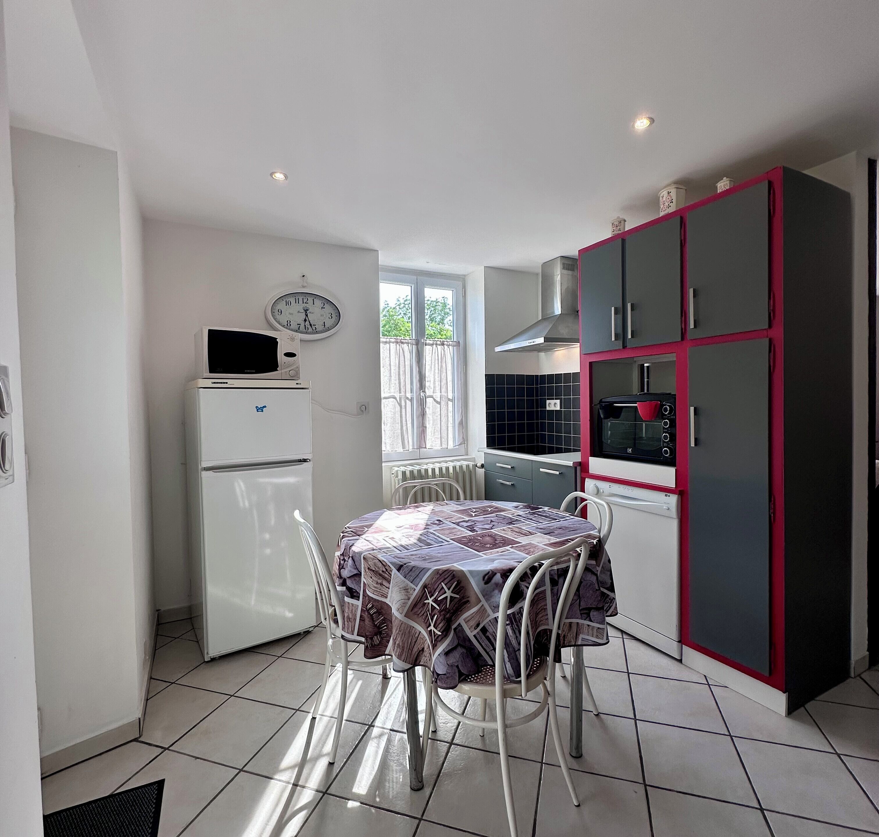 Una cocina compacta con refrigerador, microondas y horno rojo y negro. Hay una mesa de comedor con sillas y un reloj en la pared.