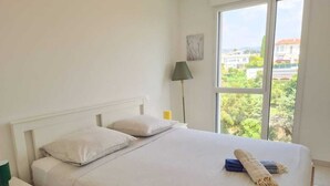 Una cama bien hecha con cabecera blanca y almohadas, una lámpara de noche y vistas a la vegetación a través de un gran ventanal.