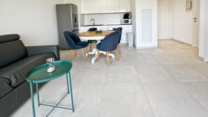 Una sala moderna con un sofá gris, una mesa de centro verde azulado y sillas azules. Se ve una cocina con armarios blancos y un refrigerador al fondo.