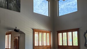 A spacious living room with a large L-shaped black leather sofa, a ceiling fan, and two large windows with wooden shutters. - Relax in the Gilbert Getaway (Gilbert)