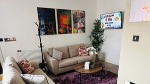 A living room with a beige sofa, a coffee table, and a purple rug. There are abstract paintings on the wall and a poster with text.