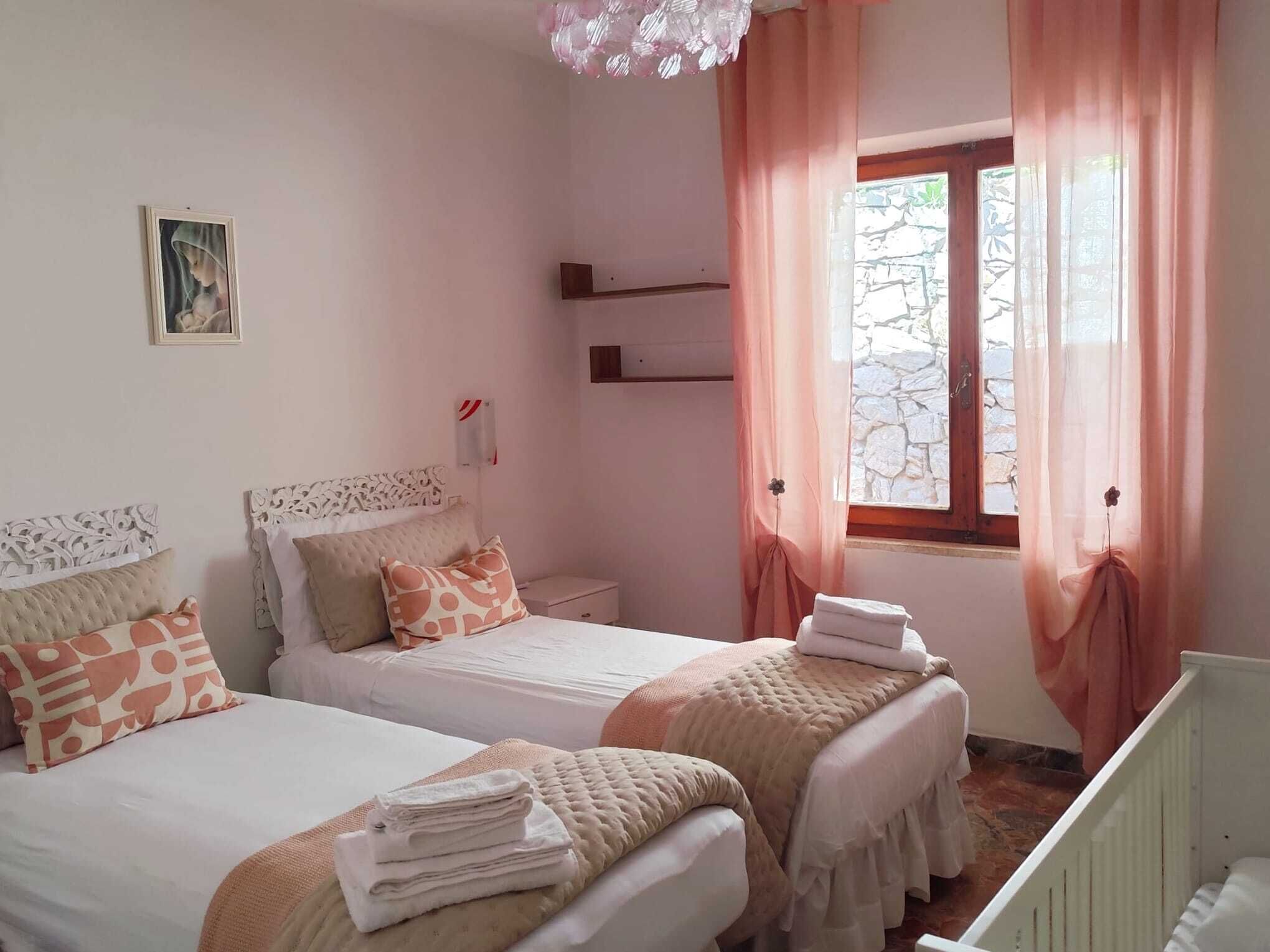 A bedroom with two beds, a window with curtains, a radiator, and a wall-mounted shelf.