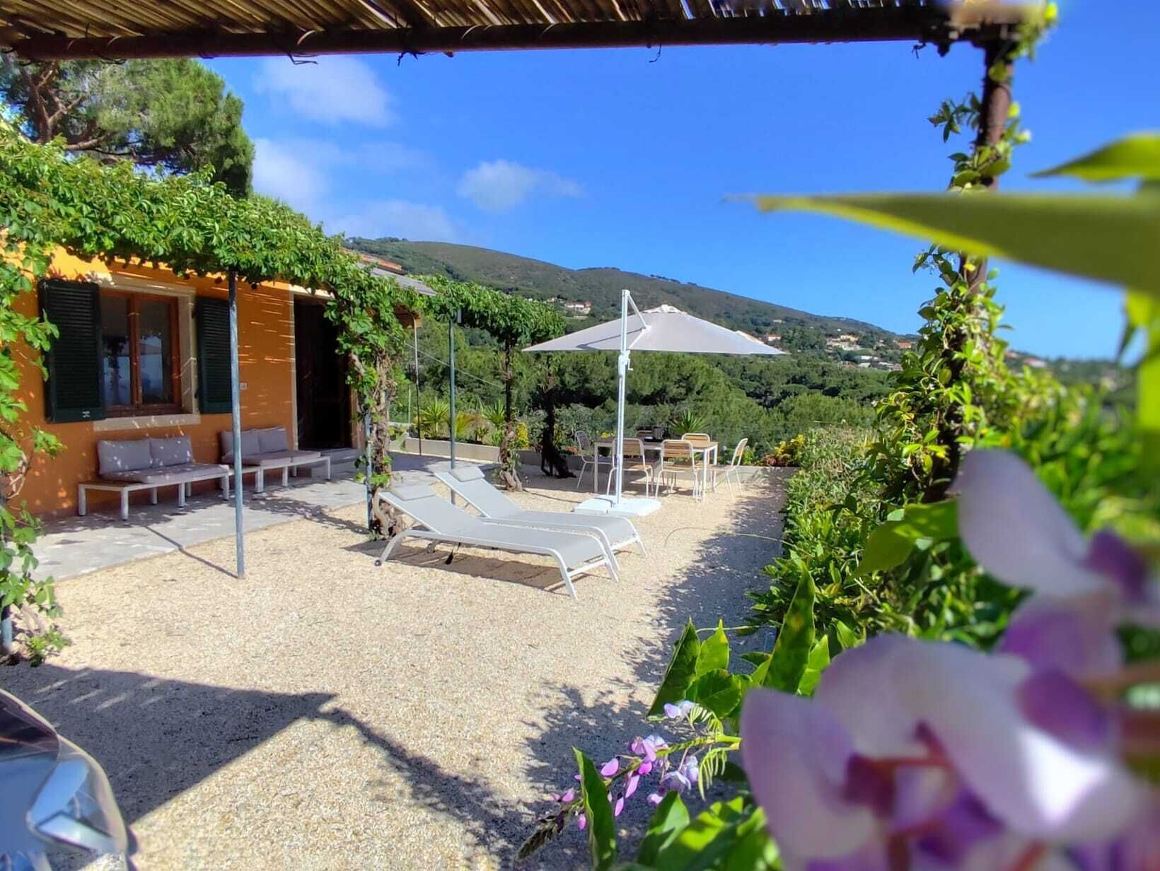 A covered outdoor seating area with lounge chairs and a table, surrounded by greenery and a view of hills.