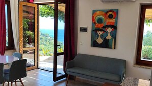 A room with a sofa, a dining table with chairs, a painting, and a view of the sea.