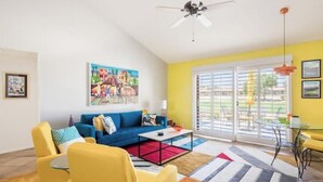 A modern living room with a blue sofa, yellow armchairs, and a coffee table. There is a colorful painting on the wall, a ceiling fan, and a glass door leading to an outdoor area.