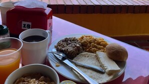 A table with breakfast items, including cereal, toast, and a plate of food, set against a backdrop of a lush garden and a lake. - Hosteria florida tropical (Olaya)