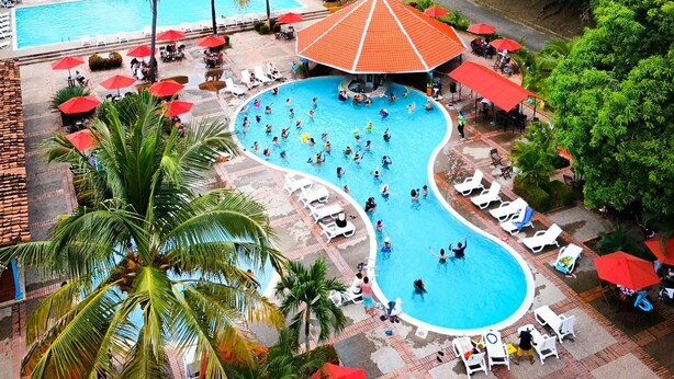 Ein Resort mit großem Pool, umgeben von Palmen und roten Sonnenschirmen, vor einer Kulisse üppiger, grüner Berge.