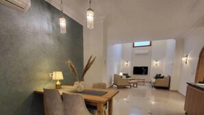 A modern living room with a dining area, featuring a sofa set, a TV mounted on the wall, and pendant lighting. - 2BRs with garden- Three Sisters Villa 3 (New Cairo City)
