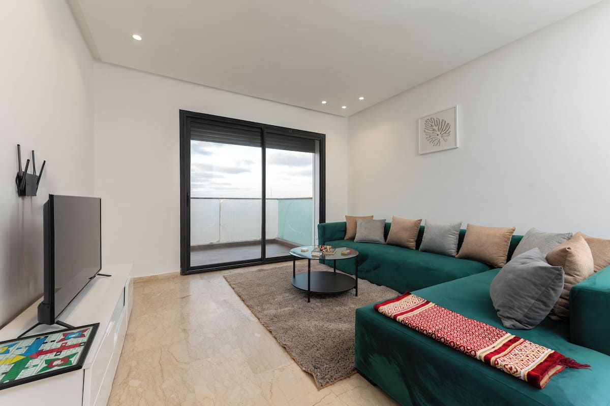 A modern living room with a green sofa, a flat-screen TV, a small coffee table, and a large window with a view of the sea.