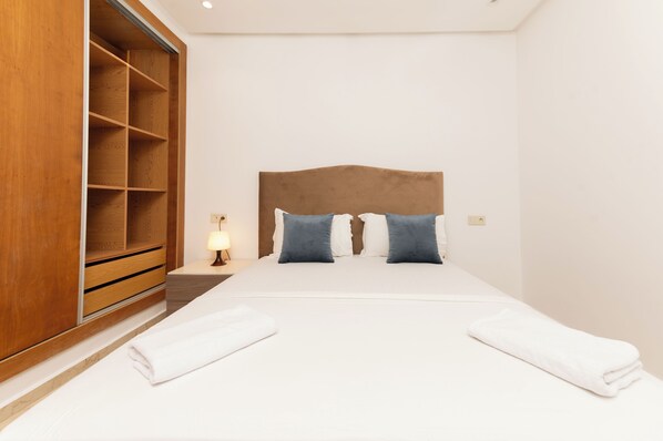 Una cama bien hecha con cabecera de madera, dos almohadas y dos toallas blancas dobladas.