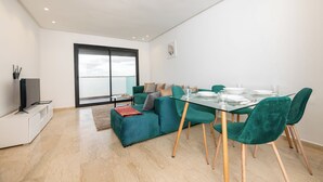 A modern living room with a teal sofa, a glass dining table with green chairs, a flat-screen TV on a white cabinet, and a large window with blinds. - Garden Bay Signature AppartHotel (Sidi Rahal)