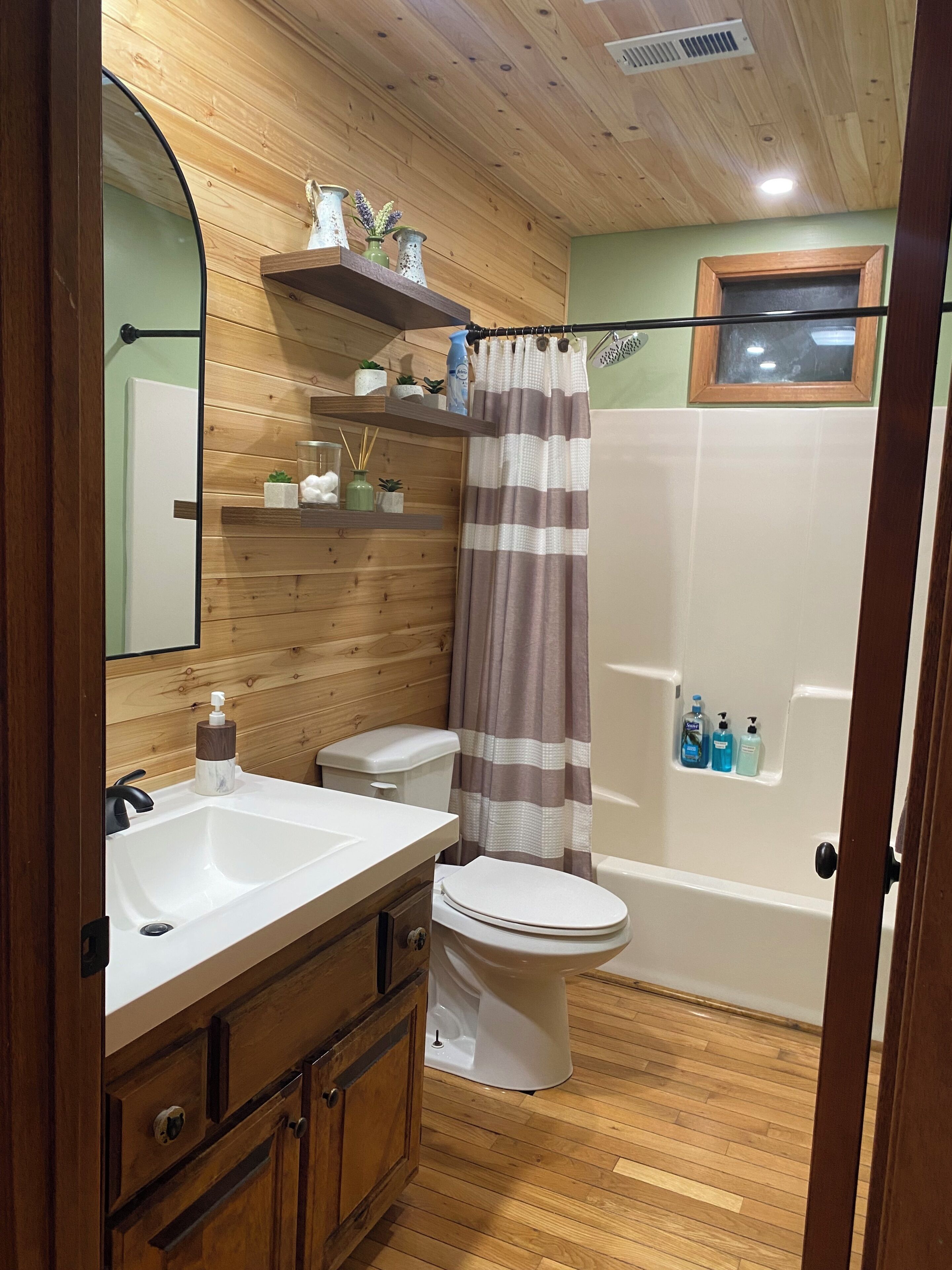 Een badkamer met houten lambrisering, een witte wastafel, een toilet, een douche met gestreept gordijn, en een klein raam boven de douche.