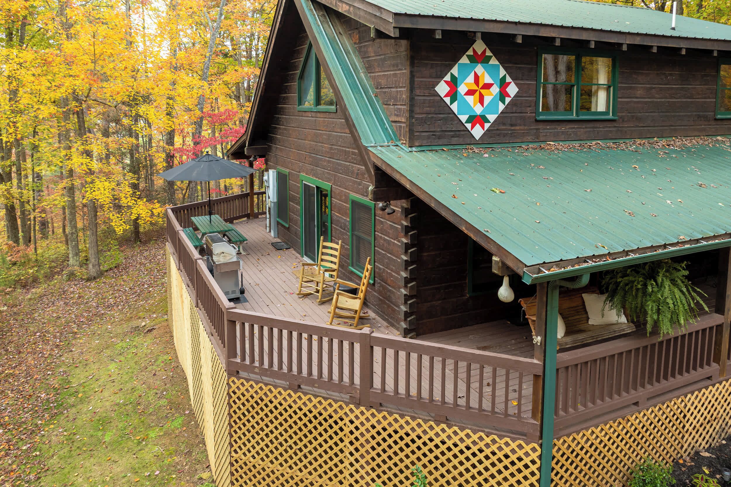 A wooden cabin with a deck, a quilt hanging on the wall, and a view of autumn trees.