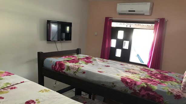 A single bed with a floral bedspread, a television mounted on the wall, and a window with pink curtains. - Hostel Rota 37 (Natal)