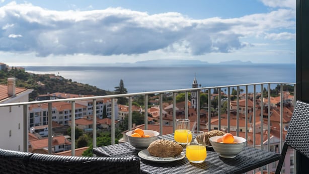 Un balcón con alimentos para el desayuno, como jugo de naranja, pan y huevos, con vistas a un pueblo costero con edificios y el mar.